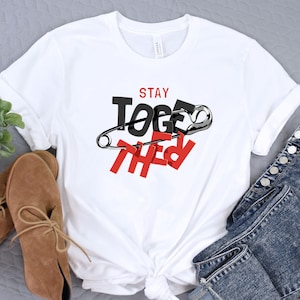 May include: White t-shirt with the words "STAY TOGETHER" in black and red, with a safety pin graphic. The shirt is styled with brown shoes and blue jeans.
