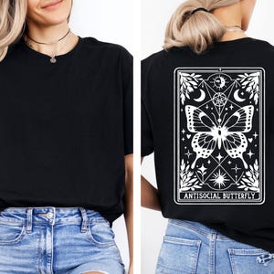 a woman wearing a black t-shirt with a graphic design on the back. The design features a butterfly and various celestial elements, including a moon and stars. The woman is also wearing blue jeans and a necklace.