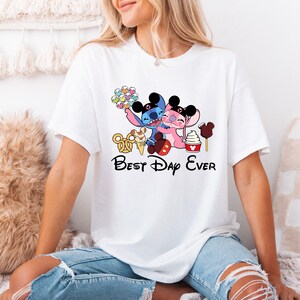 A woman wearing a white t-shirt with a graphic design featuring two cartoon characters, one pink and one blue, along with the text "Best Day Ever".