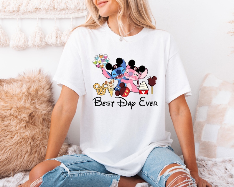 A woman wearing a white t-shirt with a graphic design featuring two cartoon characters, one pink and one blue, along with the text "Best Day Ever".