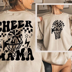 Könnte beinhalten: Ein beiger Pullover mit schwarzem Text, der "Cheer Mama" lautet, und einer Grafik eines Cheerleader-Pompons in einem Basketballkorb.