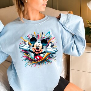 a woman sitting on a bed wearing a mickey mouse sweatshirt
