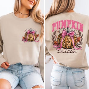 Pode incluir: Camisola bege com um design de abóbora com estampa de leopardo e um laço rosa. A frente apresenta uma versão menor do gráfico da abóbora. A parte de trás da camisola tem as palavras "PUMPKIN Season" em letras rosa acima e abaixo do gráfico da abóbora.