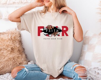 May include: Beige t-shirt with the word "FEAR" in red, with the letters torn apart to reveal the word "FAITH" and hands reaching through. The phrase "FAITH OVER FEAR" is printed below the graphic.