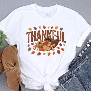 May include: White t-shirt with the word "THANKFUL" in brown letters. A cornucopia filled with pumpkins, gourds, and fall leaves is below the text. The shirt is paired with blue jeans and brown shoes.