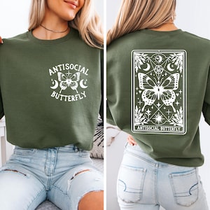 May include: Olive green sweatshirt with the text "Antisocial Butterfly" and a butterfly graphic on the front. The back features a framed butterfly design with the same text. The sweatshirt is a casual, comfortable style.