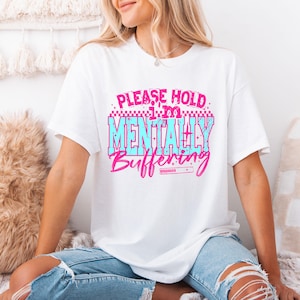 May include: White t-shirt with the text "PLEASE HOLD I'M MENTALLY BUFFERING" in pink and blue. The shirt has a crew neck and short sleeves. The design features a retro video game aesthetic.