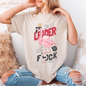 Könnte beinhalten: Beiges T-Shirt mit einem rosa Flamingo, der eine Sonnenbrille und eine Krone trägt. Der Text lautet "Yeah! Leader of the Flock" in Rot und Schwarz. Eine Sprechblase sagt "Yep! It's me."