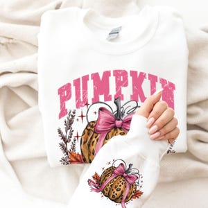 May include: White sweatshirt with a pink "PUMPKIN" graphic. The design features a leopard print pumpkin with a pink bow, surrounded by autumn leaves. The sleeve also displays the pumpkin design.