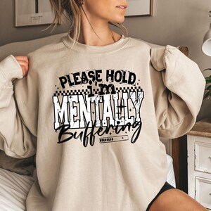 May include: Beige sweatshirt with the text "PLEASE HOLD I'M MENTALLY BUFFERING" in black and white. The text is in a bold, blocky font with a loading bar graphic. The sweatshirt is a crew neck style.
