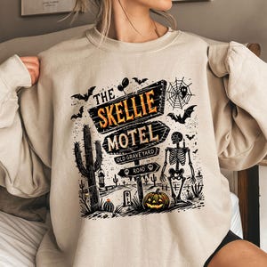 May include: Beige sweatshirt featuring a Halloween-themed graphic. The design includes a sign that reads "The Skellie Motel" with a skeleton, bats, a pumpkin, and a cactus. The words "Old Graveyard Road" are also visible.