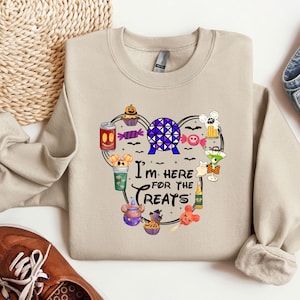 Puede incluir: Sudadera beige con cuello redondo con un gráfico colorido de golosinas y el texto "I'm here for the treats". El gráfico incluye una magdalena, caramelos y una bebida. La sudadera está doblada con zapatos marrones y vaqueros.
