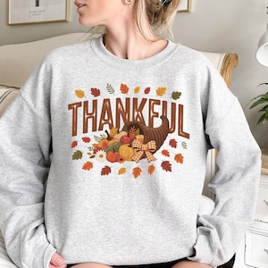 Puede incluir: Sudadera gris claro con un diseño temático de Acción de Gracias. El diseño presenta la palabra "THANKFUL" en letras marrones, una cornucopia llena de calabazas y calabacines, y hojas de otoño en tonos rojos, naranjas y amarillos.