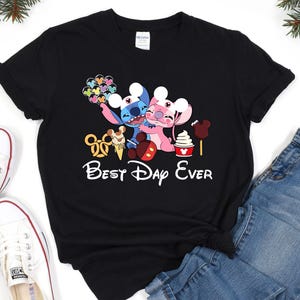 A black t-shirt with a Disney-themed graphic design, including characters from the Disney movie Lilo & Stitch, along with various Disney-related items such as a Mickey Mouse ear, a hot dog, and a cone. The text "Best Day Ever" is also present on the t-shirt.