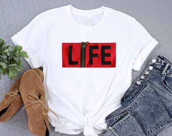 May include: White t-shirt with a red rectangle featuring the word "LIFE" in black letters. A black zipper runs vertically through the center of the rectangle. The shirt is paired with blue denim jeans and brown shoes.