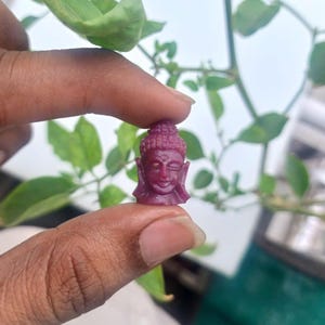 May include: A small, deep magenta-colored carved gemstone figurine depicting a Buddha head. The carving features detailed facial expressions, including closed eyes and a serene smile, with a textured top knot hairstyle and elongated earlobes.