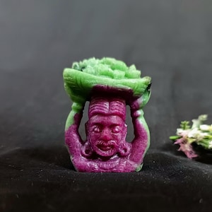 May include: A small carved figurine of a person carrying a basket of fruit on their head. The figurine is made of green and purple gemstone.