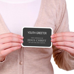 May include: A person holding a black and white name tag that reads "Youth Greeter, The Church of Jesus Christ of Latter-day Saints".