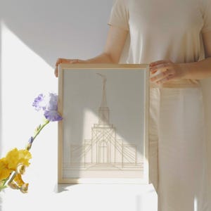 May include: A framed artwork featuring a geometric line drawing of a building with a tall spire. The artwork is in a light beige color and is set in a natural wood frame. A yellow and purple flower is in the foreground.