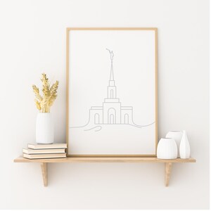 Könnte beinhalten: Ein gerahmter Druck mit einer minimalistischen Strichzeichnung eines Gebäudes mit Turm, gehalten in einer Hand. Das Kunstwerk befindet sich in einem hellen Holzrahmen und steht auf einem Holzregal mit weißen Vasen, Büchern und getrocknetem Weizen.