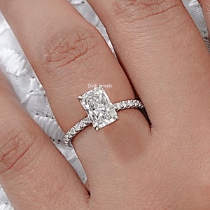 May include: A close-up of a diamond ring with a rectangular center stone. The ring is set on a band with small, sparkling stones. The ring is on a person's finger.