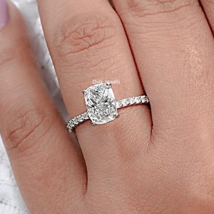 May include: A silver ring with a cushion-cut diamond set in a pave setting. The ring is on a person's finger.