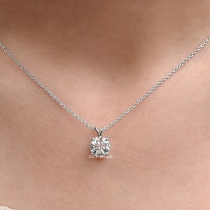 May include: A silver necklace with a square-cut, clear gemstone pendant. The pendant is suspended from a delicate chain. The necklace is displayed on a person's neck, against a neutral background. The jewelry is from the brand Dvk Jewels.