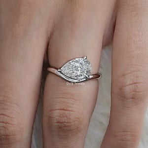 May include: A pear-shaped diamond ring with a swirling band. The ring features a large, clear diamond set in a silver or white gold band. The ring is shown on a hand with a light pink nail polish.