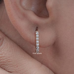 May include: A close-up of a person's ear wearing a small, gold hoop earring. The earring is lined with small, clear gemstones. The earring is a simple, elegant design. The brand name "Dvik Jewels" is visible on the earring.