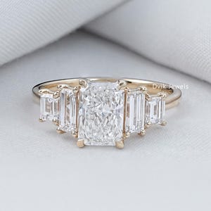 Könnte beinhalten: Ein goldener Ring mit einem großen, rechteckig geschliffenen Diamanten als Mittelstein, flankiert von kleineren Diamanten im Baguetteschliff. Der Ring ist auf einem zarten Goldband gefasst und zeigt ein klassisches und elegantes Design. Die Diamanten funkeln vor einem weichen, weißen Hintergrund.