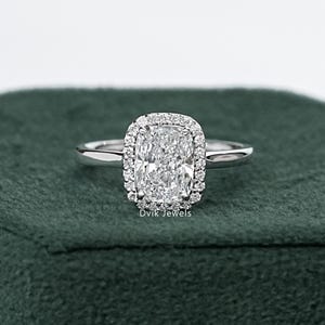 May include: A silver engagement ring with a rectangular, cushion-cut center stone surrounded by a halo of smaller diamonds. The ring is set on a dark green velvet box, creating a contrast that highlights the ring's sparkle.