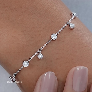 May include: A delicate silver bracelet featuring a series of round and square-cut clear gemstones. The bracelet is shown on a wrist, with the gemstones dangling from a fine chain. The bracelet is a piece of jewelry.