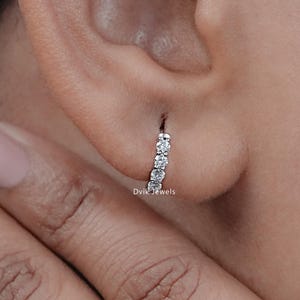 May include: A close-up of a person's ear wearing a small, silver hoop earring with a row of clear, round gemstones.
