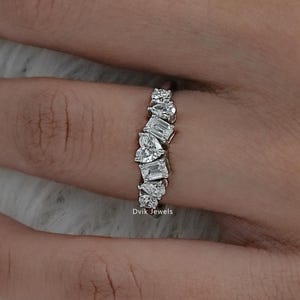 May include: A silver-toned ring featuring a cluster of multi-shaped clear stones, including emerald, pear, and heart cuts, arranged in a vertical row. The band is thin and metallic, and the text Dvik Jewels is printed near the base of the ring.