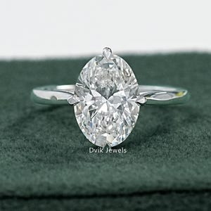 May include: A solitaire engagement ring featuring a large, oval-cut diamond set on a slender, polished silver band. The diamond sparkles brightly, reflecting light. The ring is displayed on a dark green surface, with the text "Dvik Jewels" visible.
