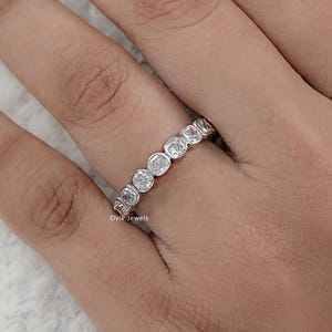 May include: A silver ring with a series of round, clear gemstones set in a bezel style. The ring is on a person's finger, with the hand and part of the arm visible. The ring is a delicate piece of jewelry.