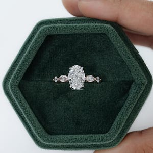 May include: An engagement ring with a large oval-cut diamond center stone and smaller accent stones on a rose gold band. The ring is displayed in a dark green velvet ring box. The ring box is in the shape of a hexagon.