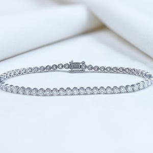 May include: A delicate tennis bracelet featuring a series of round-cut diamonds set in a silver or white gold band. The bracelet is secured with a clasp and rests on a white surface. This elegant piece of jewelry is suitable for special occasions.