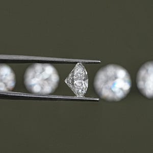 May include: A close-up of a diamond held in tweezers. The diamond is clear and sparkling. There are other diamonds out of focus in the background.
