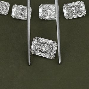 May include: A close-up of a clear, rectangular, faceted gemstone held by tweezers against a green background. The gemstone is likely a diamond.