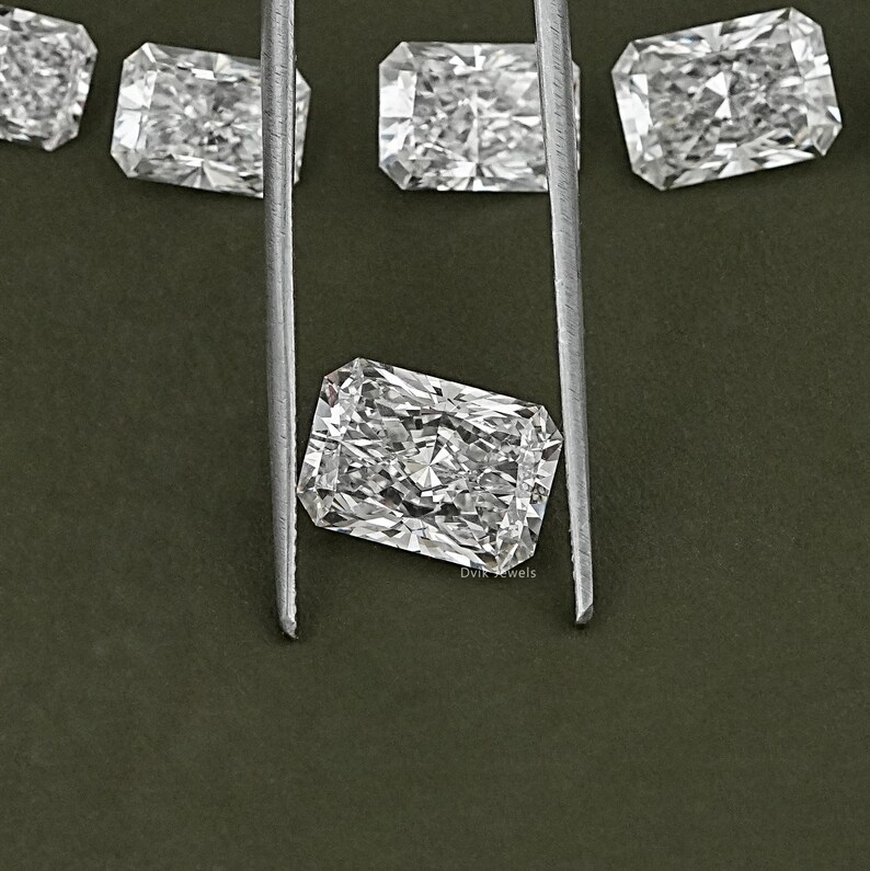 May include: A close-up of a clear, rectangular, faceted gemstone held by tweezers against a green background. The gemstone is likely a diamond.
