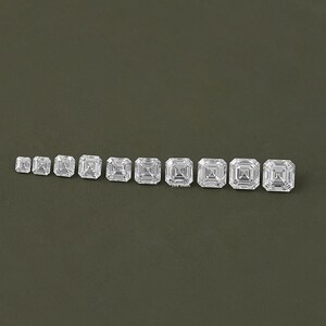 May include: A row of ten clear, square-shaped, faceted gemstones of varying sizes, arranged from smallest to largest.