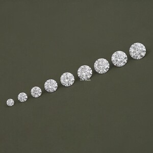 May include: A set of ten round, clear, faceted gemstones of varying sizes, arranged in a line from smallest to largest on a dark green background.