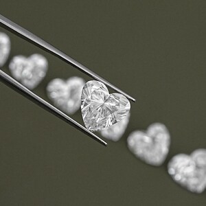 May include: A clear heart-shaped diamond gemstone held by tweezers. The diamond is surrounded by other heart-shaped diamonds, all out of focus.