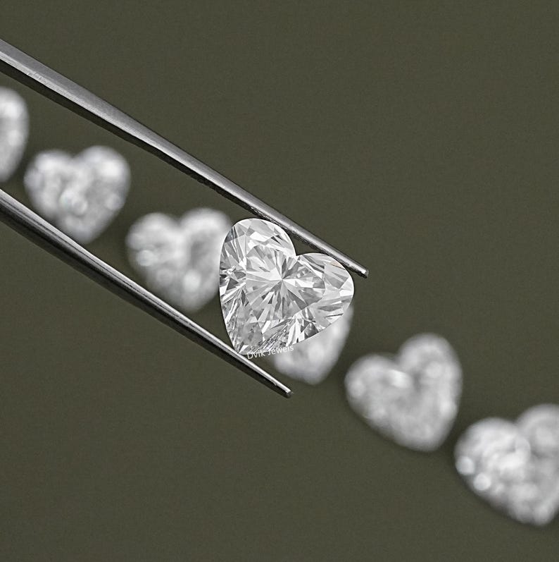 May include: A clear heart-shaped diamond gemstone held by tweezers. The diamond is surrounded by other heart-shaped diamonds, all out of focus.