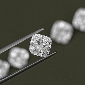 K&ouml;nnte beinhalten: Ein klarer, quadratischer Diamant, der mit einer Pinzette gehalten wird. Der Diamant ist von anderen &auml;hnlichen Edelsteinen umgeben.
