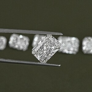 May include: A clear, rectangular, emerald cut diamond gemstone held by tweezers. The diamond is surrounded by other similar gemstones.