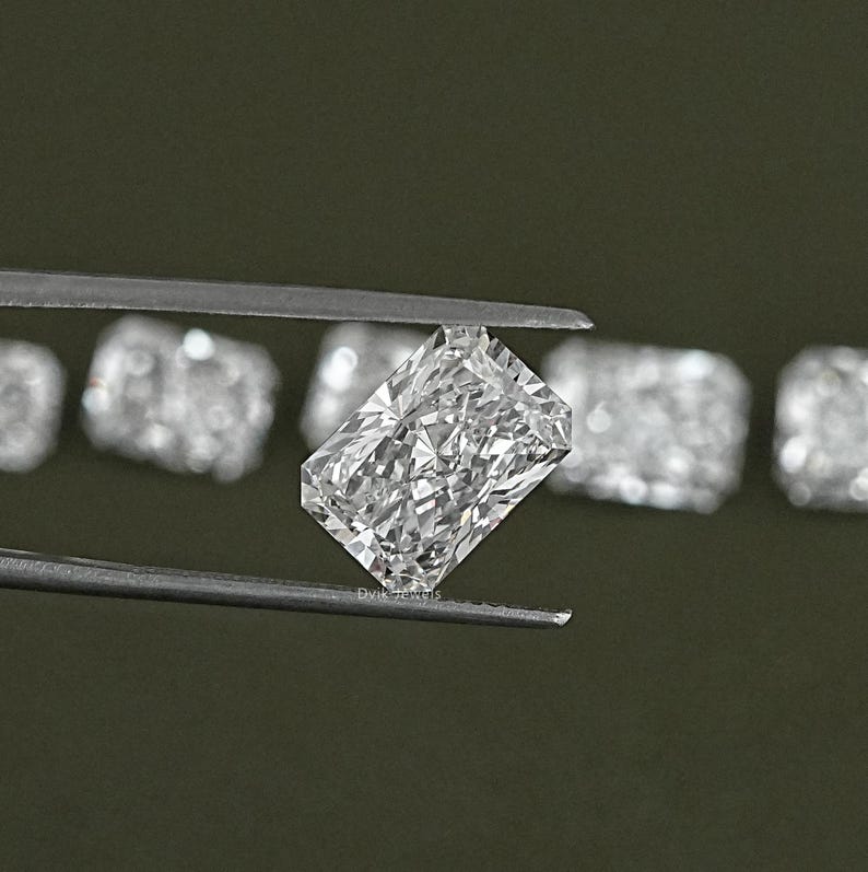 May include: A clear, rectangular, emerald cut diamond gemstone held by tweezers. The diamond is surrounded by other similar gemstones.