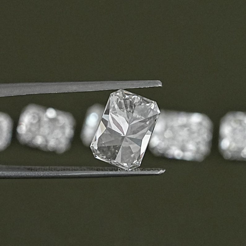 May include: A clear, rectangular, faceted gemstone held in tweezers. The gemstone is surrounded by other smaller, faceted gemstones.