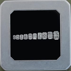 May include: A collection of radiant-cut diamonds of varying sizes, arranged in a row against a black background. The diamonds are displayed within a silver-colored frame, showcasing their brilliance and clarity. The diamonds are all rectangular in shape.